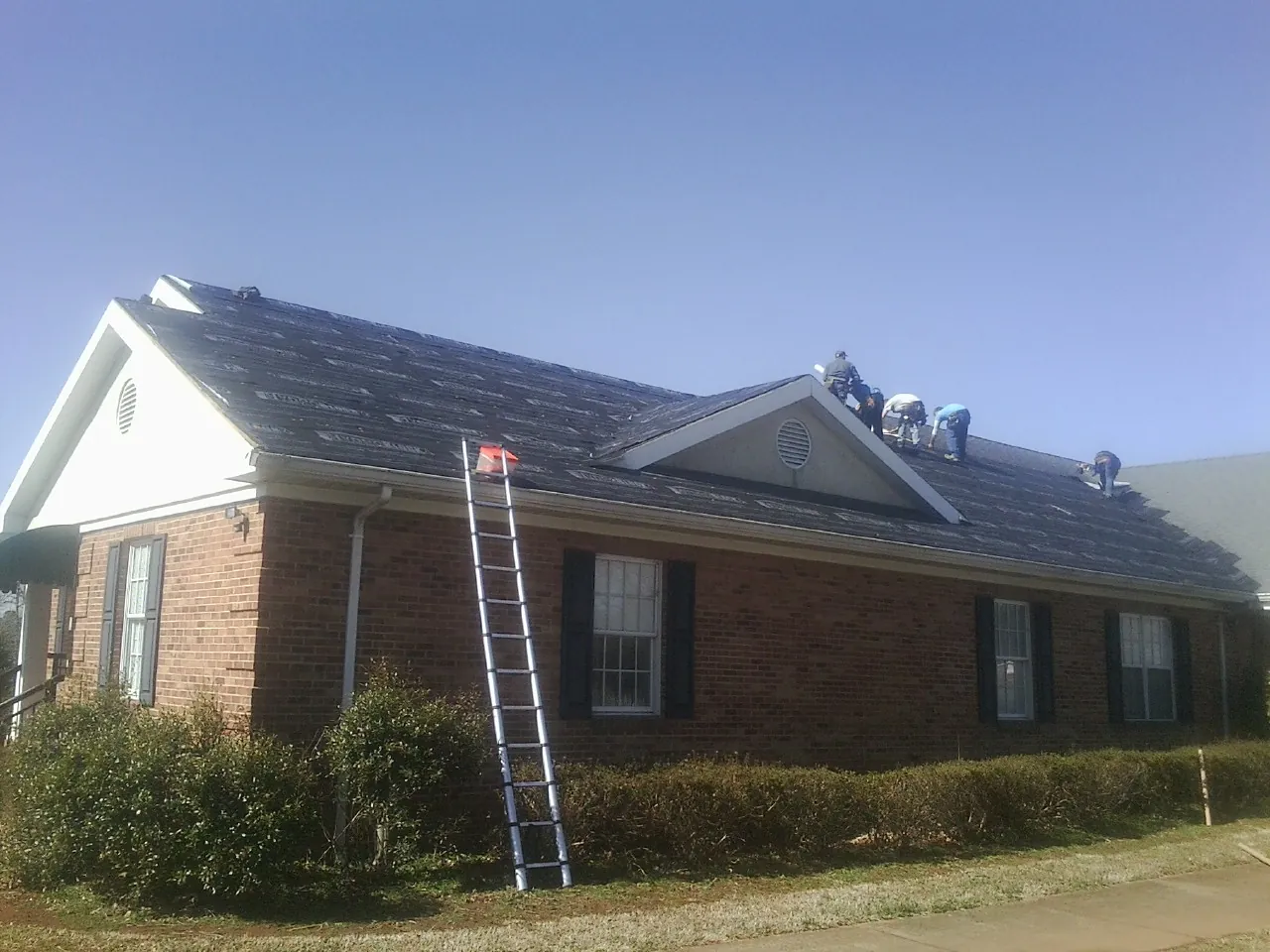 Crew tearing shingles off a brick ranch roof with a ladder set — Emergency Roof Repair work by our crew in Mc Graw