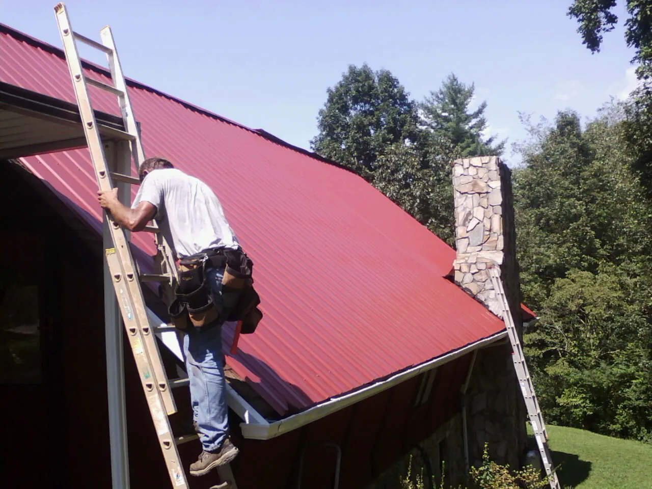 Roofer on a ladder beside a red metal roof and stone chimney — Soffit & Fascia Repair by Roofing Brothers in Mc Graw